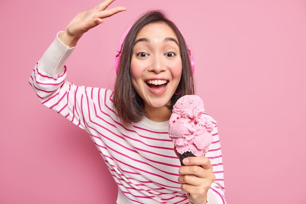 Indoor shot of brunette woman with eastern appearance dances carefree raises arm smiles gladfully eats delicious ice cream listens music wears casual striped jumper isolated over pink wall.