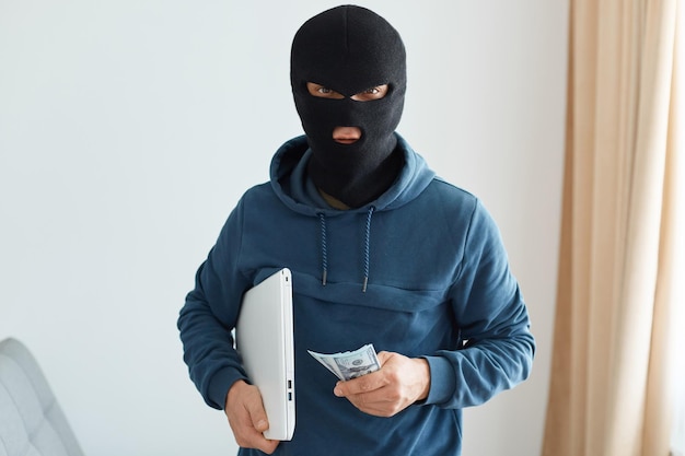 Indoor shot of burglar robbed the house and leaves with the stolen laptop and money, male burglar wearing dark blue hoodie and black mask, performed illegal actions.