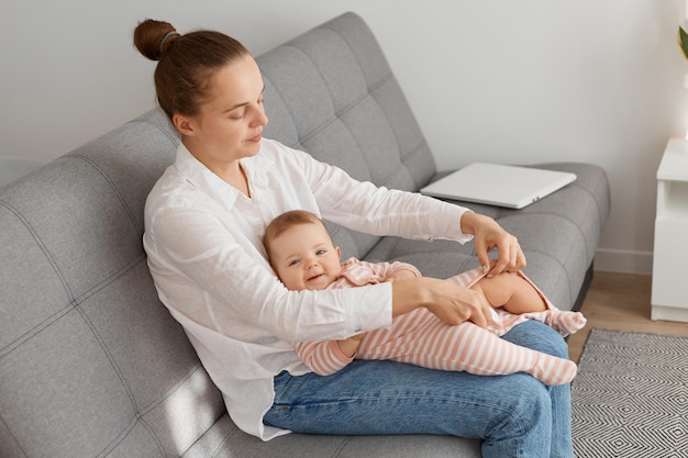 Indoor shot of caring mother wearing casual clothing, having hair bun, sitting on sofa and dressing her infant baby daughter, preparing for going for a walk.