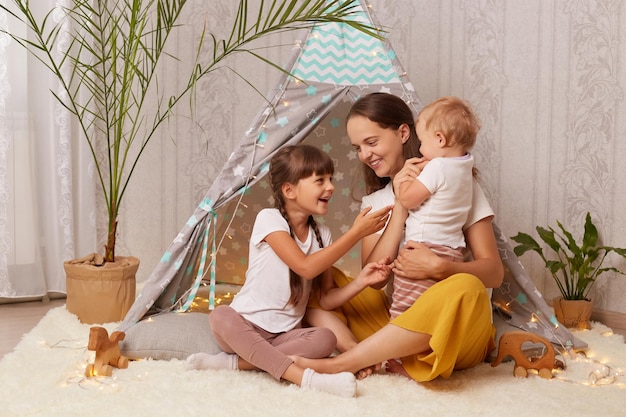 Indoor shot of Caucasian attractive woman with her children sitting together teepee tent mom playing with daughters spending weekend with kids while playing in wigwam