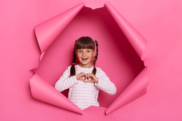 Indoor shot of charming excited little girl wearing striped shirt posing in pink paper torn standing looking at camera showing heart gesture with her palms