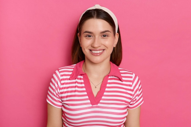 Indoor shot of cheerful charming young girl with hair band wearing striped casual t shirt looking at camera with toothy smile standing isolated pink background expressing positive emotions