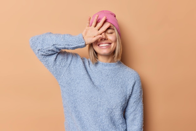 Indoor shot of cheerful pretty teenage girl keeps eyes closed covers face with hand smiles gladfully shows white teeth doesnt hide emotions dressed in casual jumper isolated over brown background.