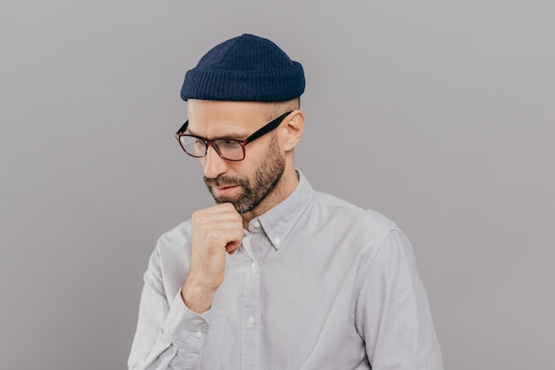 Indoor shot of concentrated man holds chin and focused down being in deep in thoughts thinks about innovative idea for his research has clever expression wears glasses and white formal shirt