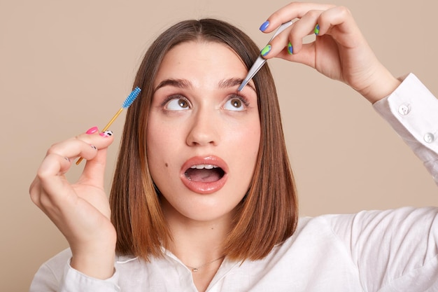 Indoor shot of concentrated woman with tweezers and eyebrow brush plucking brows hair using metal forceps for eyebrows posing with open mouth looking up isolated over beige background