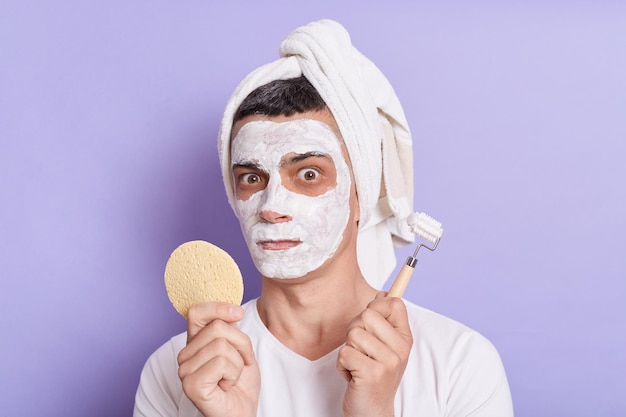 Indoor shot of confused Caucasian man with mask for skin wrapped in towel standing with massage roller and sponge in hands isolated over violet background