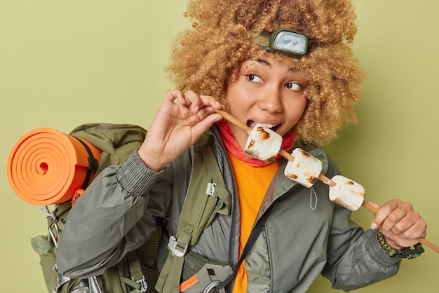 Indoor shot of curly haired woman camper eats roasted marshmallow wears headlamp and jacket carries rucksack full of necessary equipment has picnic isolated over green background Recreation concept