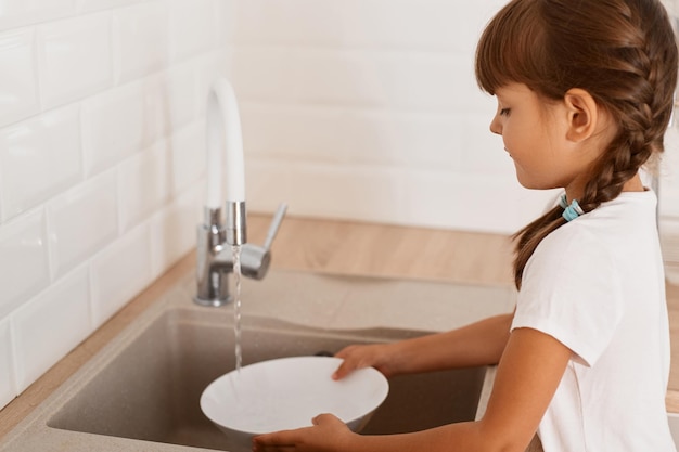 Indoor shot of cute charming little girl washing white plate in the kitchen dark haired female kid in white t shirt doing household chores helping her mother