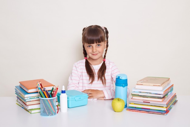 Indoor shot of cute smart little girl with braids sitting at desk isolated over white background being surrounded with school supplies looking at camera with serious expressing being ready to study