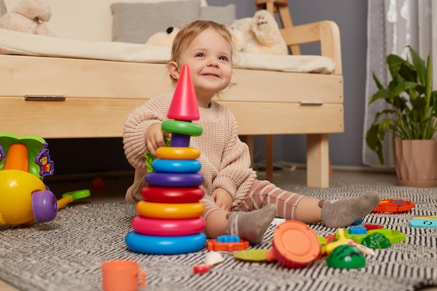 Indoor shot of cute smiling happy cheerful baby girl sitting on floor with her toys playing with colorful pyramid plays educational games alone at home