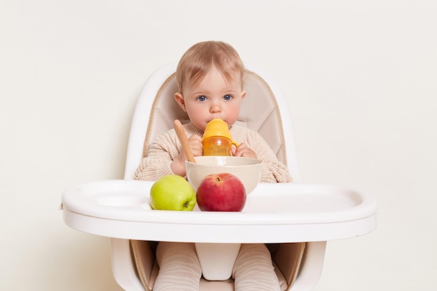 Indoor shot of cute thirsty baby wearing beige sweater sitting in a child's chair and drinking water from yellow bottle isolated on a white background