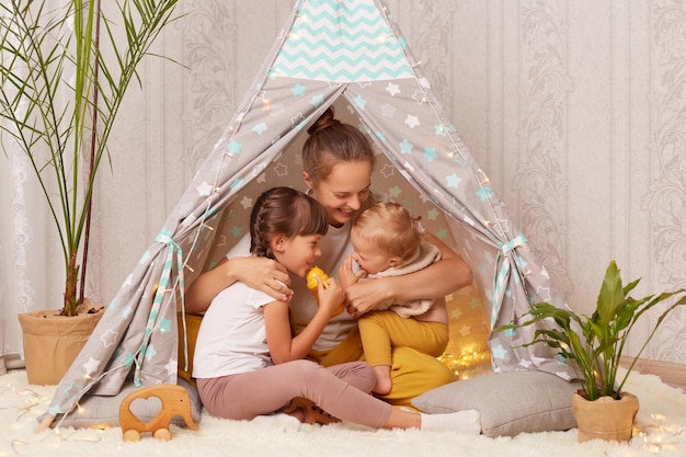 Indoor shot of dark haired Caucasian woman with two daughters sitting together in wigwam hugging with love and laughing playing with pleasure at home