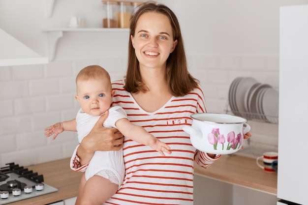 Indoor shot of dark haired woman and small child in the kitchen at home female in striped shirt holding baby in hands and pot cooking with her kid expressing happiness