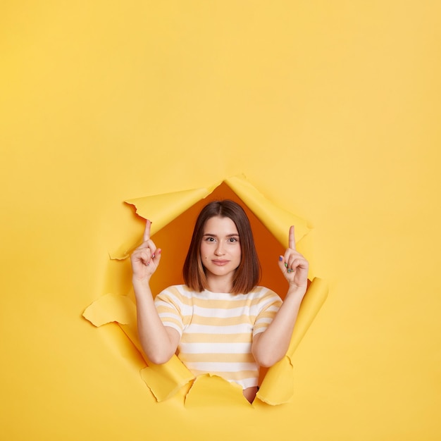 Indoor shot of dark haired young adult woman wearing striped t shirt looking through yellow paper torn pointing fingers up looks at camera copy space for advertisement