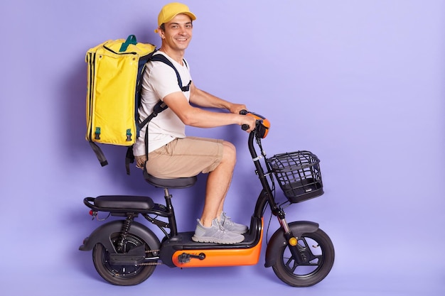 Indoor shot of delivery man wearing with thermo backpack delivering food on electric scooter isolated over purple background riding to client with fast food order looking at camera