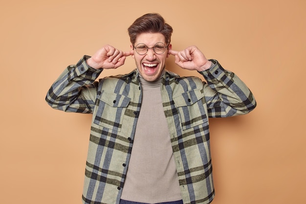 Indoor shot of emotional young European man plugs ears exclaims loudly keeps mouth opened avoids bothering noise wears round spectacles and casual checkered shirt isolated over brown background