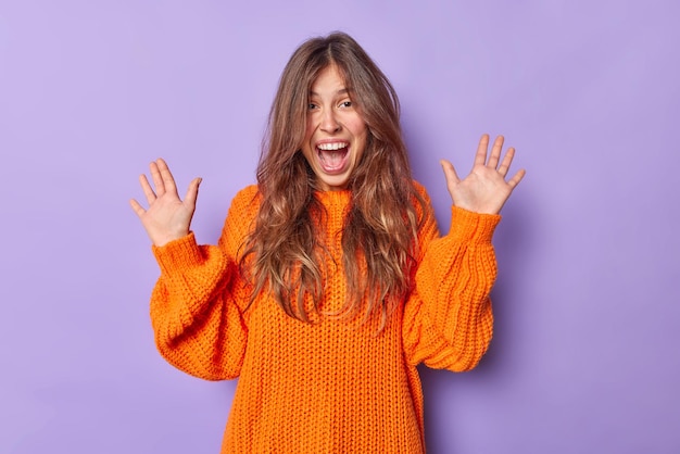 Indoor shot of emotional young woman exclaims loudly keeps palms raised up reacts happily on awesome news dressed in loose knitted sweater isolated over purple background. Happiness concept.