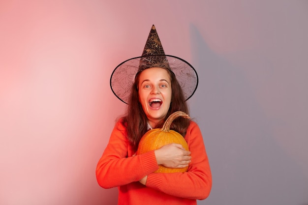 Indoor shot of excited amazed Caucasian woman dressed in witch hat holding orange pumpkin isolated on neon light background extremely happy female celebrating halloween