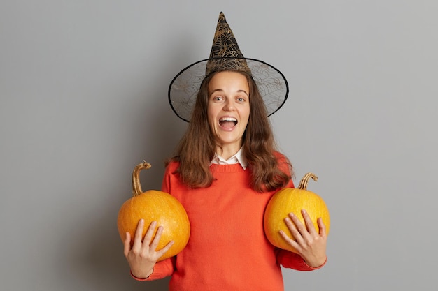 Indoor shot of excited amazed Caucasian woman wearing sweater and witch hat standing with orange pumpkin in hands yelling happily while celebrating Halloween posing isolated over gray background