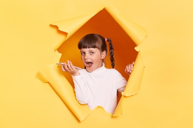 Indoor shot of excited amazed little girl with pigtail breaking through yellow paper and holding cell phone recording voice message expressing positive emotions