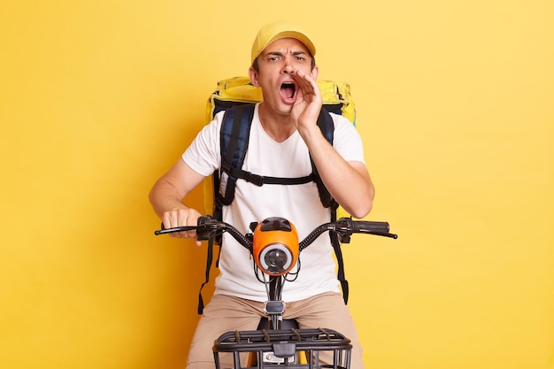 Indoor shot of excited deliveryman on bicycle wearing white Tshirt and cap isolated over yellow background screaming announcement loud keeps hand near mouth delivering orders