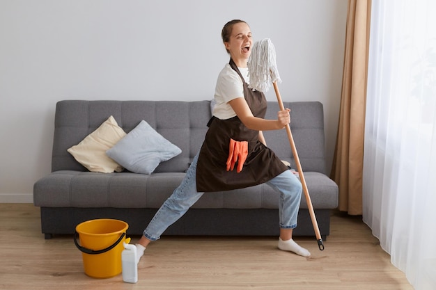 Indoor shot of excited young female wearing jeans and brown apron cleaning her home singing at mop using tool as like microphone and having fun in living room