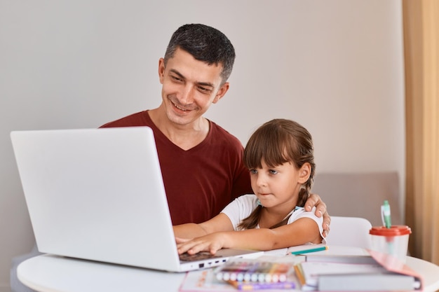 Indoor shot of father wearing maroon t shirt doing homework with daughter having online lesson on laptop dad and small girl learn at home have web class on computer