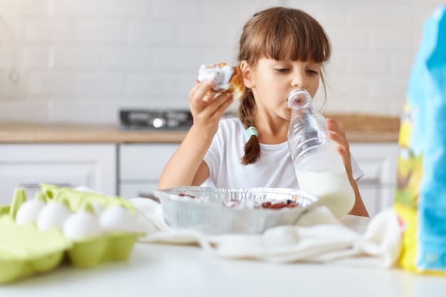 Indoor shot of female kid wearing white casual t shirt, having dark hair and pigtails eating homemade croissant and drinking milk from the bottle, enjoying yummy baking.