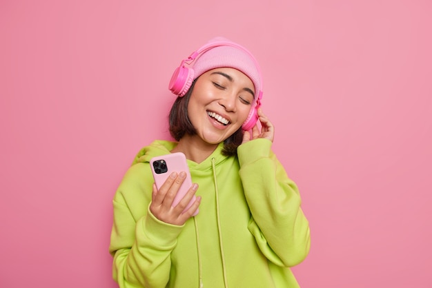 Indoor shot of happy Asian woman tilts head listens favorite music from playlist wears stereo headphones holds mobile phone wears casual green sweatshirt and hat isolated over pink wall