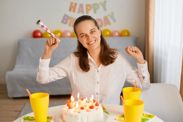 Indoor shot of happy attractive woman with dark hair sitting at table with party tube in hands looking at camera being ready to blow candles and making wish celebrating her birthday