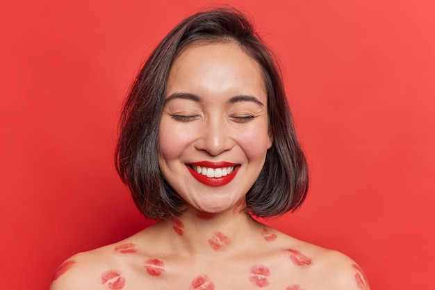 Indoor shot of happy feminine lady with eastern appearance smiles toothily keeps eyes closed wears red lipstick poses shirtless has lips traces on body isolated over vivid studio wall