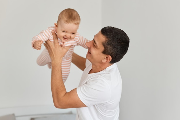 Indoor shot of happy handsome brunette man wearing white casual t shirt holding his infant daughter, posing at home in light room, kid flying like plane in father's hands.