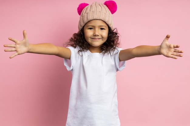 Indoor shot of happy little girl with dark kinky hair, wearing white t shirt and knitted hat, giving hug, stretching both hands, smiling, isolated. Child joyfully meeting parents.