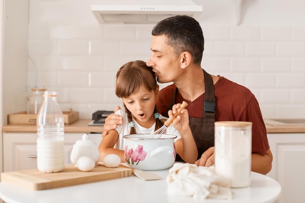 Indoor shot of happy lovely family posing in white kitchen, father kissing his daughter while making pie or cake together, father and child preparing tasty surprise for mother.