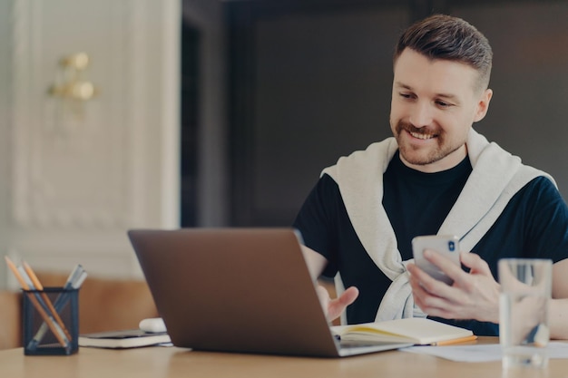 Indoor shot of happy male freelancer uses modern gadgets searches information on laptop computer hods modern smartphone poses at desktop against cozy interior installsnew application uses internet