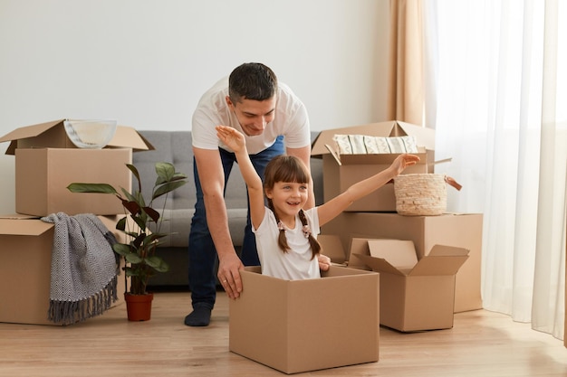 Indoor shot of happy positive brunette father wearing white t shirts paying with her daughter during relocation to a new house child with raised arms posing in carton box celebrating relocation