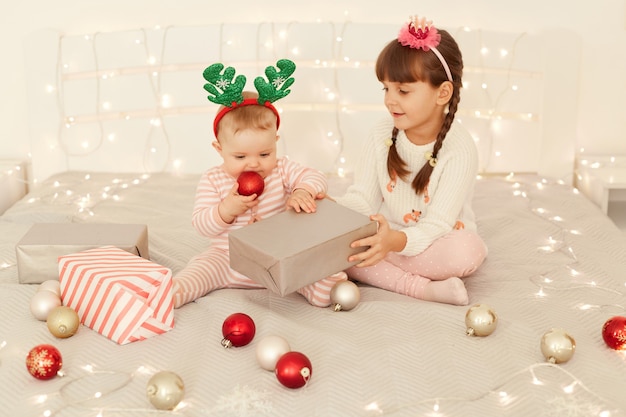 Indoor shot of happy positive children sitting on bed with Christmas baubles and garland lights, elder girl giving present box to infant baby, New year eve.