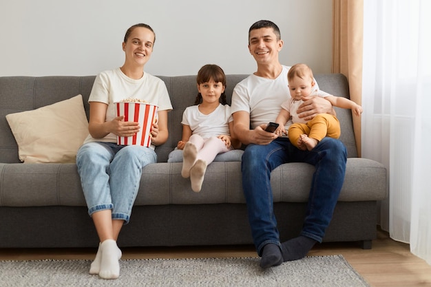 Indoor shot of happy positive family wearing casual white tshirts sitting on sofa watching tv show or movie together with children enjoying spending free time smiling happily