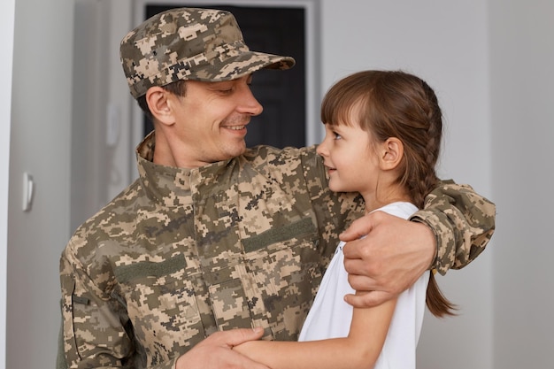 Indoor shot of happy positives soldier in camouflage uniform an cap hugging his daughter returning home from war posing at home with door and white wall on background