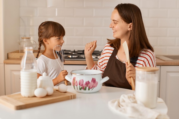 Indoor shot of happy satisfied young adult woman cooking with her daughter in the kitchen, having fun together with her charming kid, expressing positive emotions.