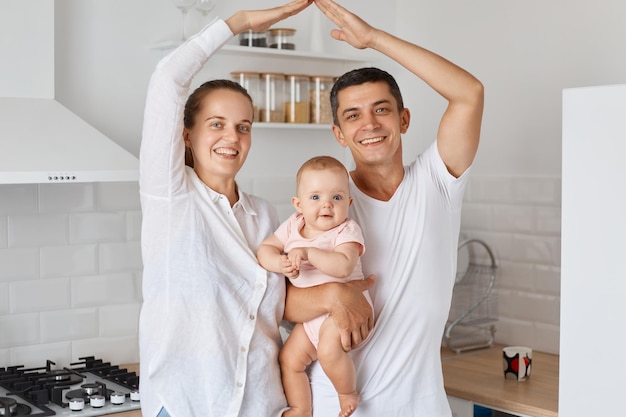 Indoor shot of happy smiling couple standing with baby daughter and making roof with hands above their heads feels in safety together at home expressing positive emotions