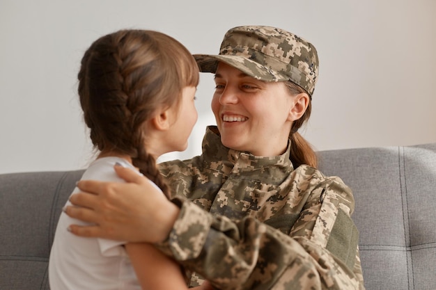 Indoor shot of happy young adult military woman wearing camouflage uniform and cap posing with her daughter looking at each other with love and hugging