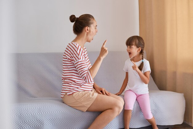 Photo indoor shot of hard working woman speech therapist working with little girl at home female wearing striped shirt and shorts sitting on sofa with child