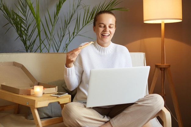 Indoor shot of joyful optimistic beautiful woman wearing white jumper sitting on cough and working on laptop or watching film eating fast food enjoying pizza