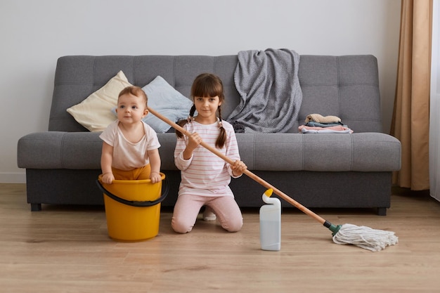 Indoor shot of little girl with mop in hands and her infant sister doing household the living room together having fun while tidying up the apartment baby sitting in bucket