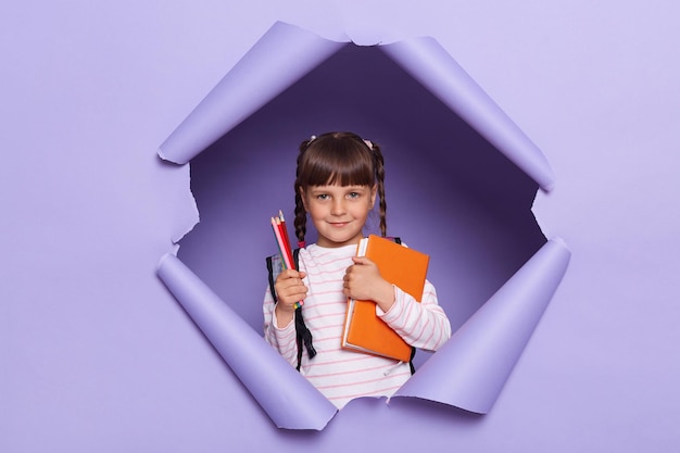 Indoor shot of little girl with pigtails wearing striped shirt breaks through purple paper background holding book and pens being ready going to school and study waiting the start of the lesson