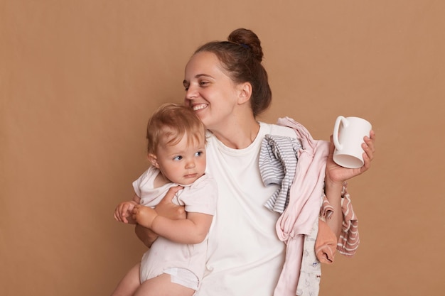 Indoor shot of lovely loving young mother with bun hairstyle hugging her toddler daughter holding cup in hands expressing happiness posing isolated over brown background