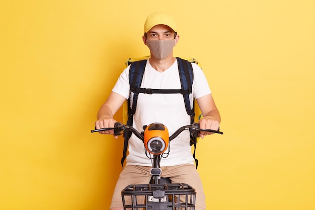 Indoor shot of man courier riding cycle wearing white t shirt cap and protective mask courier delivery fast delivering during quarantine looking at camera isolated over yellow background