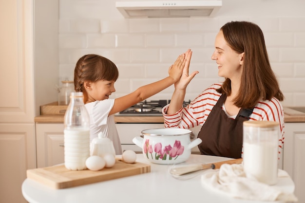 Indoor shot of mother and her cute charming little daughter playing and giving five to each other while baking in kitchen at home surrounded with bake ingredients