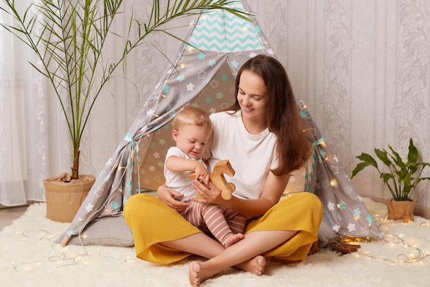 Indoor shot of mother with baby girl playing at home in wigwam siting on the floor and smiling happily spending time together expressing positive e motions